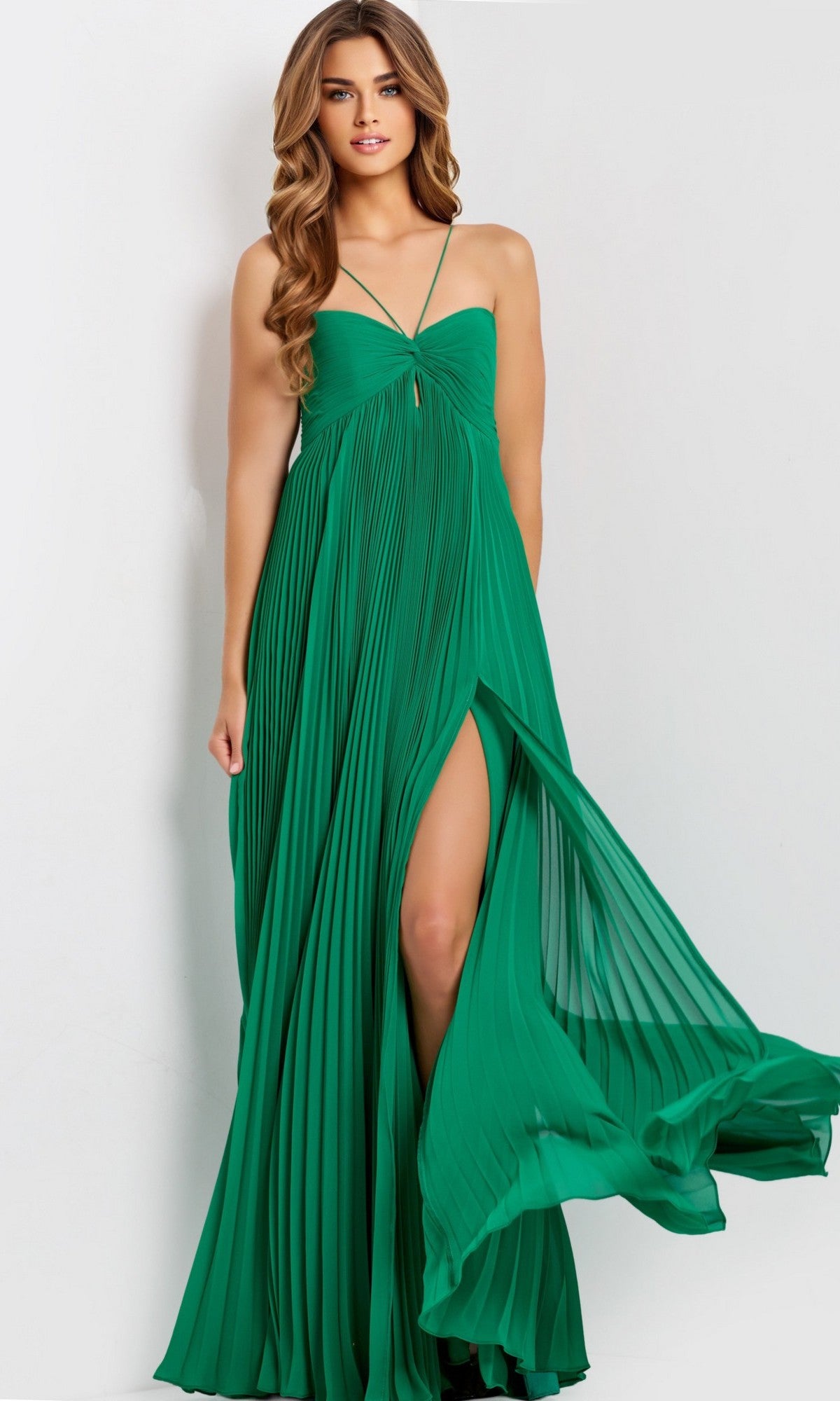 A woman stands against a plain light background wearing the Jovani 47820, a sleeveless green long prom dress with pleats and a thigh-high leg slit.
